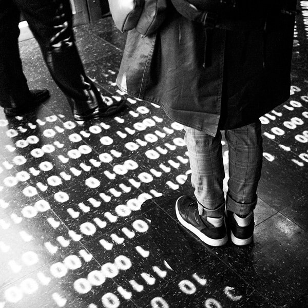 Photo en noir et blanc de deux personnes dont on ne voit que le bas du corps sur une projection binaire de 0 et de 1