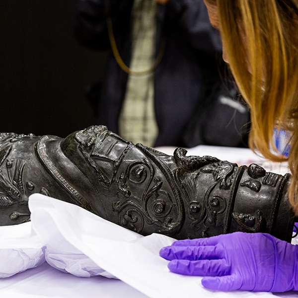 Un femme aux cheveux roux portant des gants mauves est penchée sur une jambière en bronze de gladiateur.