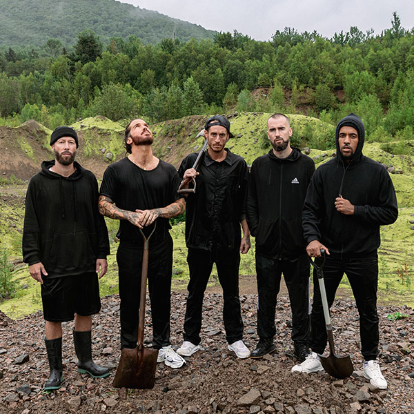 photo du groupe de musique Alaclair Ensemble. Les membres, vêtus de noir et tenant des pelles pour creuser le sol, sont photographiés sur une butte de terre avec en arrière-plan la forêt et la montagne